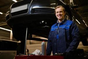 professionell porträtt av en stilig caucasian man, bil mekaniker, tekniker, bil ingenjör stående nära en låda med uppsättning av verktyg för reparation bilar i de reparera affär garage foto