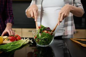 fokus på gravid kvinnas händer innehav trä- skedar och blandning Ingredienser i en glas skål, framställning utsökt friska sallad för middag i de kök ö Nästa till henne Make hackning grönsaker foto