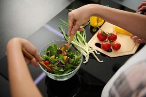 topp se av kvinnas händer innehav trä- skedar och blandning Ingredienser i en glas skål, framställning utsökt friska sallad för middag i de kök ö Nästa till henne Make hackning grönsaker foto