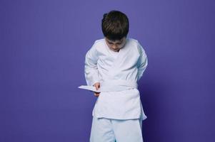 självsäker caucasian skola ålder pojke sätter på vit kimono, ties en bälte runt om hans midja, redo för bekämpa sporter. aikido kämpe. orientalisk krigisk konst foto