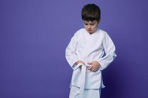 horisontell porträtt av en självsäker Tonårs pojke aikido brottare kvitt vit bälte av hans kimono, isolerat över violett bakgrund med kopia ad Plats foto