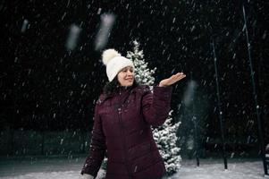 Lycklig kvinna fångst snöflingor på henne hand medan gående på stad gata, på snöig natt. glad jul Lycklig ny år foto