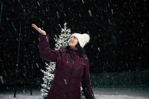 härlig kvinna fångst snöflingor på henne hand medan gående ner de stad gata på en snöig kväll. vinter. snöfall foto