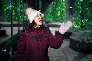skön kvinna fångster snöflingor, åtnjuter gående ner de stad gata, lättade förbi kransar på en snöig vinter- natt foto