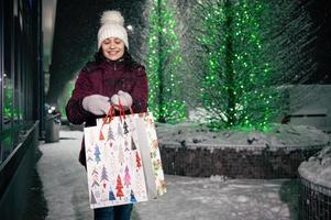 multietnisk kvinna promenader med handla påsar på de gata upplyst förbi kransar på snöig vinter- natt. glad jul foto