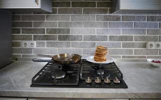 främre se av nyligen bakad hemlagad pannkakor på vit tallrik, stål fräsning panorera och spatel på en svart spis i de Hem kök. skrava tisdag begrepp foto