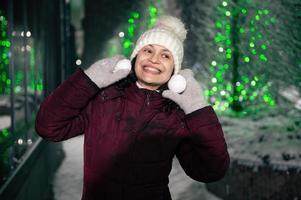 härlig kvinna i värma kläder, leende en skön toothy leende medan spelar med snöbollar på snö täckt gata foto