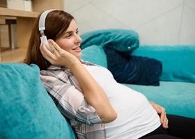 skön caucasian kvinna i förväntan av en bebis vilar på moderskap lämna, försiktigt strök henne gravid mage och njuter lyssnande till klassisk musik på hörlurar på Hem, liggande ner på en soffa foto