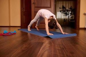 förtjusande aktiva sportig unge flicka stretching henne kropp i nedåt vänd hund utgör, praktiserande yoga i mysigt Hem interiör foto