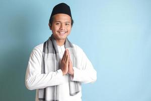 porträtt av asiatisk muslim man i vit koko skjorta med kalott som visar be om ursäkt och Välkommen hand gest. isolerat bild på blå bakgrund foto