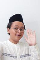 porträtt av leende ung asiatisk muslim man tar bild av han själv eller selfie, ordspråk Hej och vinka hans hand. skärm fånga av en telefon. isolerat bild på vit bakgrund foto