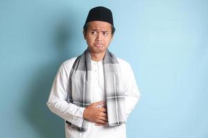 porträtt av ung asiatisk muslim man känsla smärta i hans mage. magont begrepp. isolerat bild på blå bakgrund foto