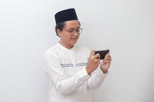 porträtt av ung asiatisk muslim man höjning hans näve, fira vinnande spel eller få Bra Nyheter på hans mobil telefon. isolerat bild på vit bakgrund foto
