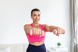 inhemsk Träning med vikter. positiv svart lady håller på med övningar med hantlar, förstärkning henne kropp på Hem. leende ung kvinna arbetssätt på henne biceps muskler, vistas friska foto