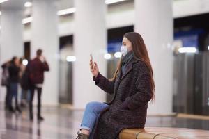 en kvinna i en medicinsk ansiktsmask väntar på ett tåg och håller en smartphone foto