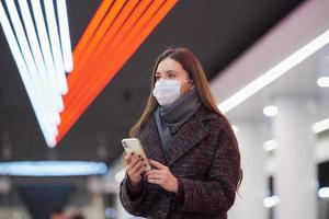 en kvinna i en medicinsk ansiktsmask väntar på ett tåg och håller en smartphone foto