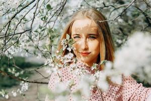 blond flicka på en vår promenad i de trädgård med körsbär blommar. kvinna porträtt, närbild. en flicka i en rosa polka punkt klänning. foto