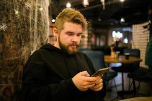 stilig koncentrerad kille framställning pengar transaktion via Ansökan på mobil telefon med 4g internet Sammanträde på Kafé i gata kväll, allvarlig tusenåriga man läsning text meddelande på smartphone foto