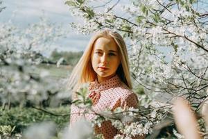 blond flicka på en vår promenad i de trädgård med körsbär blommar. kvinna porträtt, närbild. en flicka i en rosa polka punkt klänning. foto