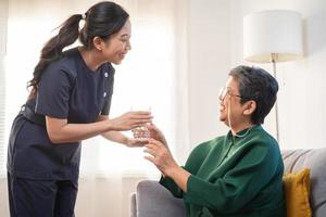 asiatisk ung kvinna vårdgivare, vårdare stödjande äldre, åldrig patient på Hem, dricka vatten efter ta medicin, bota eller behandling sjukdom av mormor efter pensionering, hjälp Stöd. foto