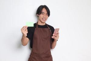 porträtt av attraktiv asiatisk Barista man i brun förkläde som visar mall av kreditera kort och pekande på Det. isolerat bild på vit bakgrund foto