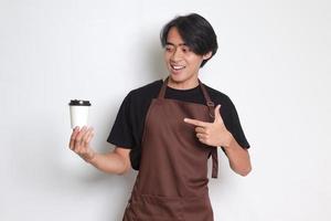 porträtt av attraktiv asiatisk Barista man i brun förkläde innehav ta bort papper kaffe kopp och pekande på Det. isolerat bild på vit bakgrund foto