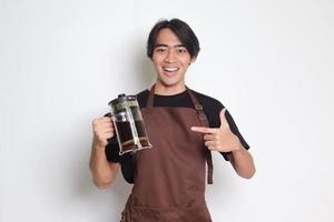 porträtt av attraktiv asiatisk Barista man i brun förkläde pekande med finger och som visar franska Tryck kaffe tillverkare. isolerat bild på vit bakgrund foto