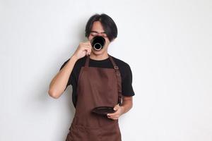 porträtt av attraktiv asiatisk Barista man i brun förkläde dricka en kopp av kaffe medan innehav de fat. isolerat bild på vit bakgrund foto