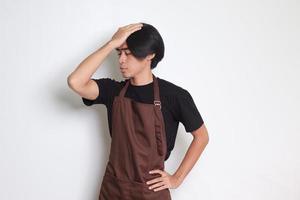 porträtt av attraktiv asiatisk Barista man i brun förkläde innehav hand på panna, glömmer något, och beklagar han själv. isolerat bild på vit bakgrund foto
