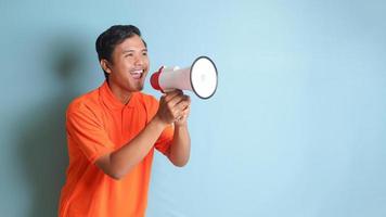porträtt av attraktiv asiatisk man i orange skjorta tala högre använder sig av megafon, främja produkt. reklam begrepp. isolerat bild på blå bakgrund foto