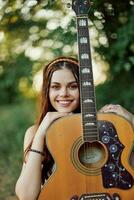 ung hippie kvinna med eco bild leende och ser in i de kamera med gitarr i hand i natur på en resa foto