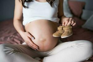 gravid kvinna avkopplande på Hem. hon är Sammanträde på säng och innehav bebis skor. foto