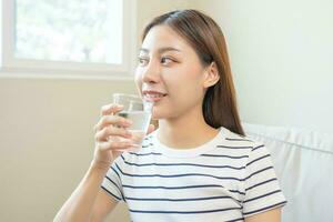 Lycklig skön, asiatisk ung kvinna, flicka dricka, smutta färsk glas av vatten för hydratisering av kropp, innehav transparent glas i henne hand, törstig på Hem. hälsa vård, friska livsstil begrepp. foto