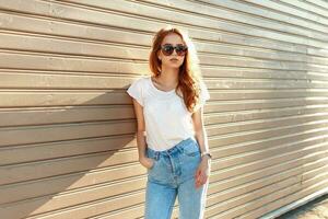 skön eleganta flicka i en årgång blå jeans och vit t-shirt stående nära en trä- vägg. foto