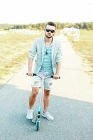 stilig ung hipster kille i en eleganta jacka i trasig shorts ridning en skoter. foto