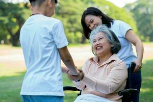 glad senior asiatisk mormor använder rullstol med sin dotter och barnbarn i parken, barnbarn kom för att besöka äldre mormor och hålla handen. begreppet lycklig familj, bra relation tillsammans foto