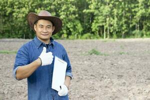 asiatisk man agronom bär hatt, blå skjorta, innehar papper urklipp, tummen upp, stå på lantbruk landa. begrepp, lantbruk forskning , undersökning och studie till utveckla jord kvalitet för växande gröda. foto