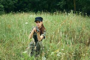 militär kvinna Sammanträde på de gräs med vapen i hand svart keps grön löv foto