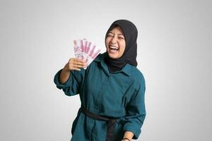 porträtt av upphetsad asiatisk hijab kvinna i tillfällig utrusta som visar ett hundra tusen rupiah. finansiell och besparingar begrepp. isolerat bild på vit bakgrund foto