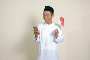 porträtt av attraktiv asiatisk muslim man i vit skjorta med kalott innehav indonesien flagga medan som visar mobil telefon. isolerat bild på grå bakgrund foto