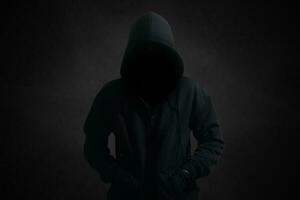 mystisk man bär svart luvtröja stående mot mörk bakgrund. hacker, brottslighet, och cyber säkerhet begrepp. foto