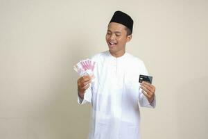 porträtt av attraktiv asiatisk muslim man i vit skjorta med kalott innehav ett hundra tusen rupiah och presenter kreditera kort. isolerat bild på grå bakgrund foto