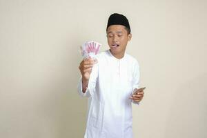 porträtt av attraktiv asiatisk muslim man i vit skjorta som visar ett hundra tusen rupiah medan använder sig av mobil telefon. finansiell och besparingar begrepp. isolerat bild på grå bakgrund foto