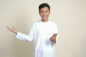 porträtt av attraktiv asiatisk muslim man i vit skjorta innehav mobil telefon med leende uttryck på ansikte medan pekande finger till de sida. reklam begrepp. isolerat bild på grå bakgrund foto