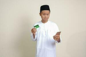 porträtt av attraktiv asiatisk muslim man i vit skjorta med kalott känsla ledsen och besviken, ser på hans mobil telefon medan innehav kreditera kort. isolerat bild på grå bakgrund foto