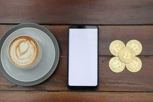 topp se av tom skärm mobil telefon med en lugg av bitcoin och kopp av kaffe på trä- tabell. kryptovaluta handel begrepp foto