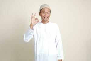 porträtt av attraktiv asiatisk muslim man i vit skjorta med kalott som visar ok hand gest och leende ser på kamera. reklam begrepp. isolerat bild på grå bakgrund foto