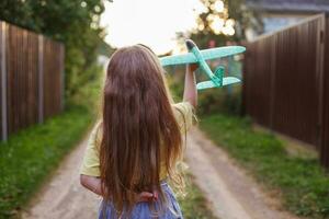 Lycklig barn flicka med lång blond hår spelar med leksak flygplan utomhus- på solnedgång foto