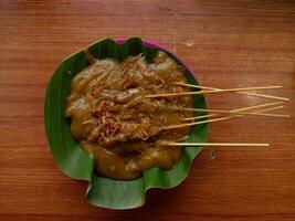 satay padang eller kallad sate padang är kryddad kyckling satay från padang, väst sumatra. eras med kryddad curry sås och ris kaka, tjäna på tallrik plast med banan blad. topp se på trä bakgrund foto