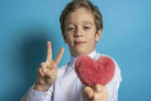 en söt caucasian pojke i en vit skjorta innehar en fluffig röd hjärta i hans händer och visar en tecken av frihet foto